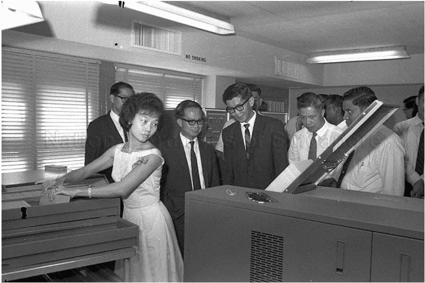 Early computer in Singapore, female punch card operator surrounded by government officials