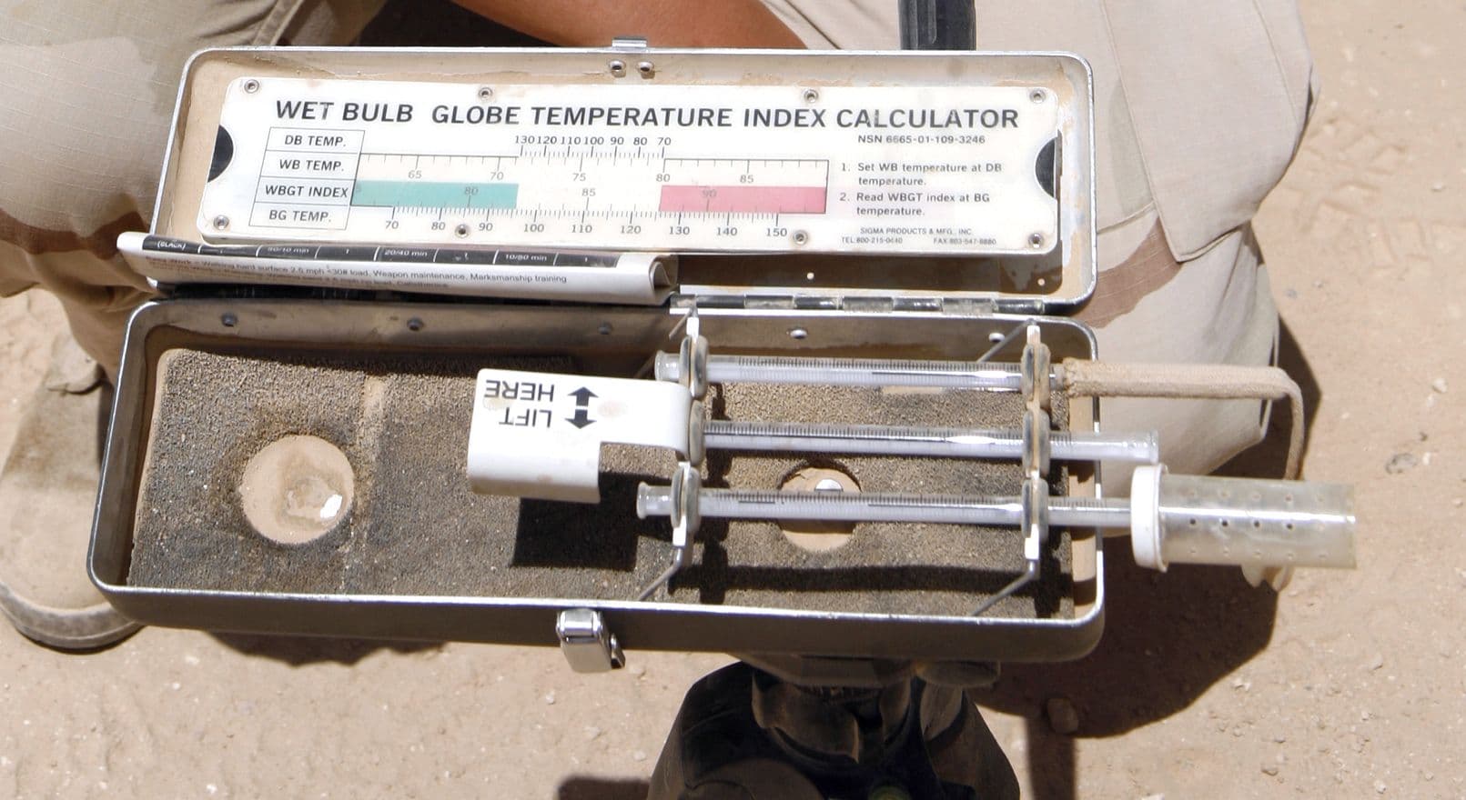 An open field kit labelled “Wet Bulb Globe Temperature Index Calculator”, holding three thermometers and a conversion chart, held over sandy ground.
