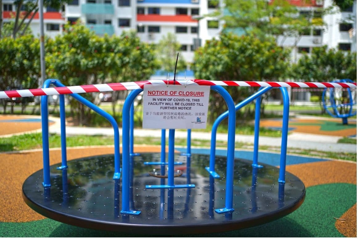 A playground roundabout in a Singapore housing estate, taped off with red-and-white barrier tape and a bilingual “Notice of Closure — in view of Covid-19” sign.